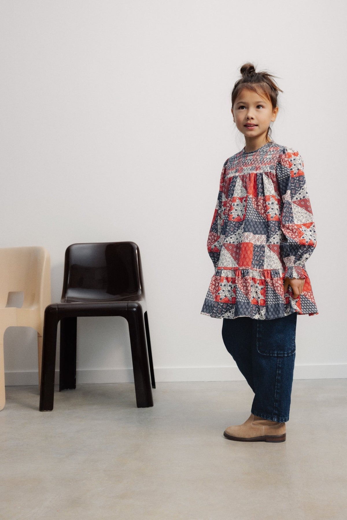 A young child standing and wearing The Quill Dress by The New Society with dark pants and boots, next to a black chair in a minimalist room.