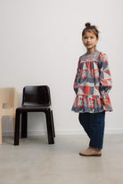A young child standing and wearing The Quill Dress by The New Society with dark pants and boots, next to a black chair in a minimalist room.