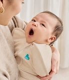 Baby wearing a beige wrap with a visible brand logo, held by an adult against a neutral background