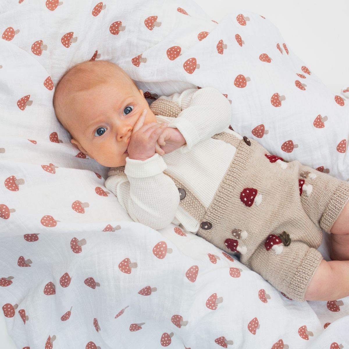 Baby lying on a white blanket with red strawberry pattern, wearing a beige outfit with red strawberries.