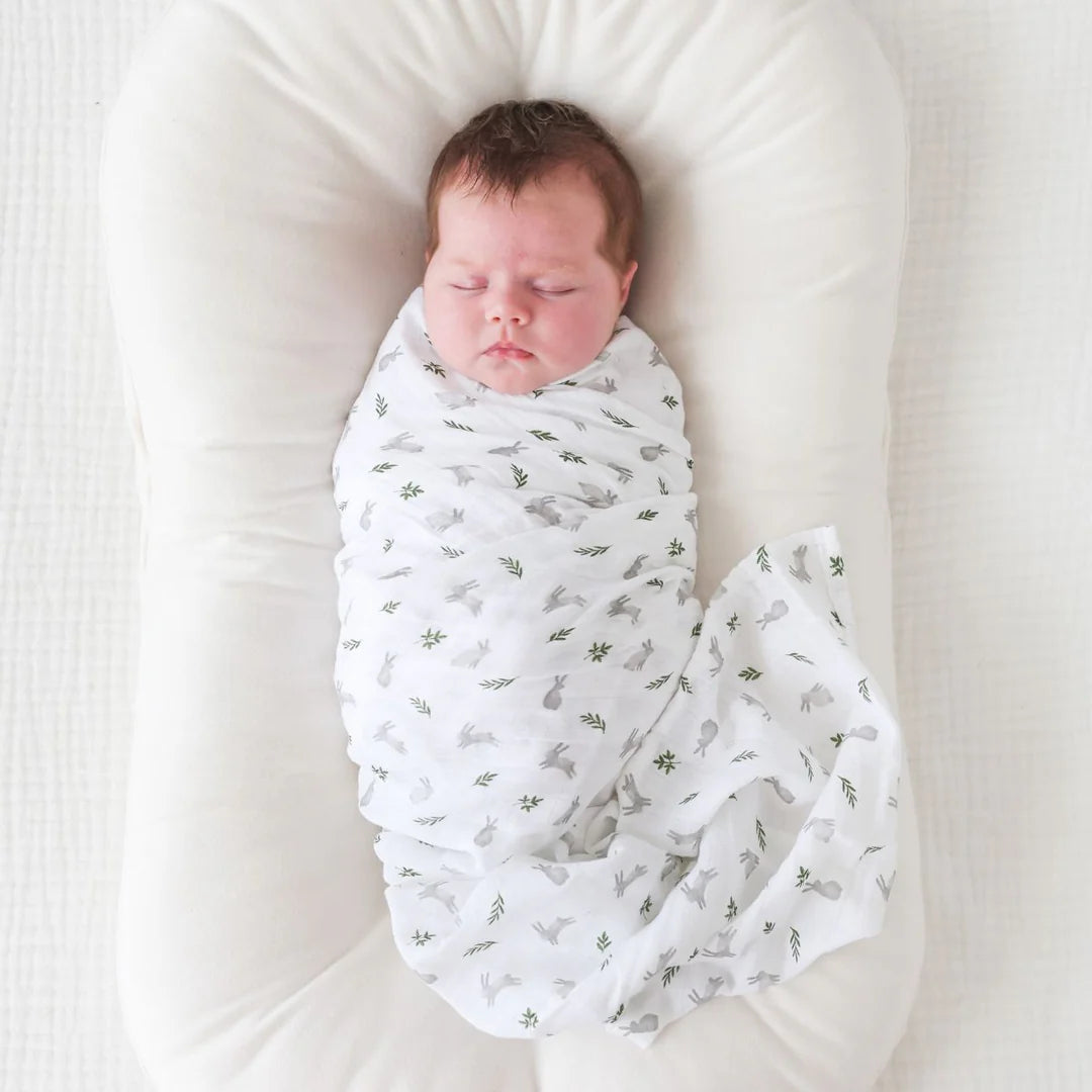 A sleeping newborn baby peacefully swaddled in a white muslin blanket with a subtle bunny and fern pattern, resting inside a white infant lounger.