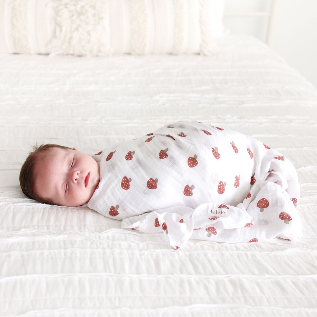 Newborn baby swaddled in a white blanket with red patterns on a white bed.