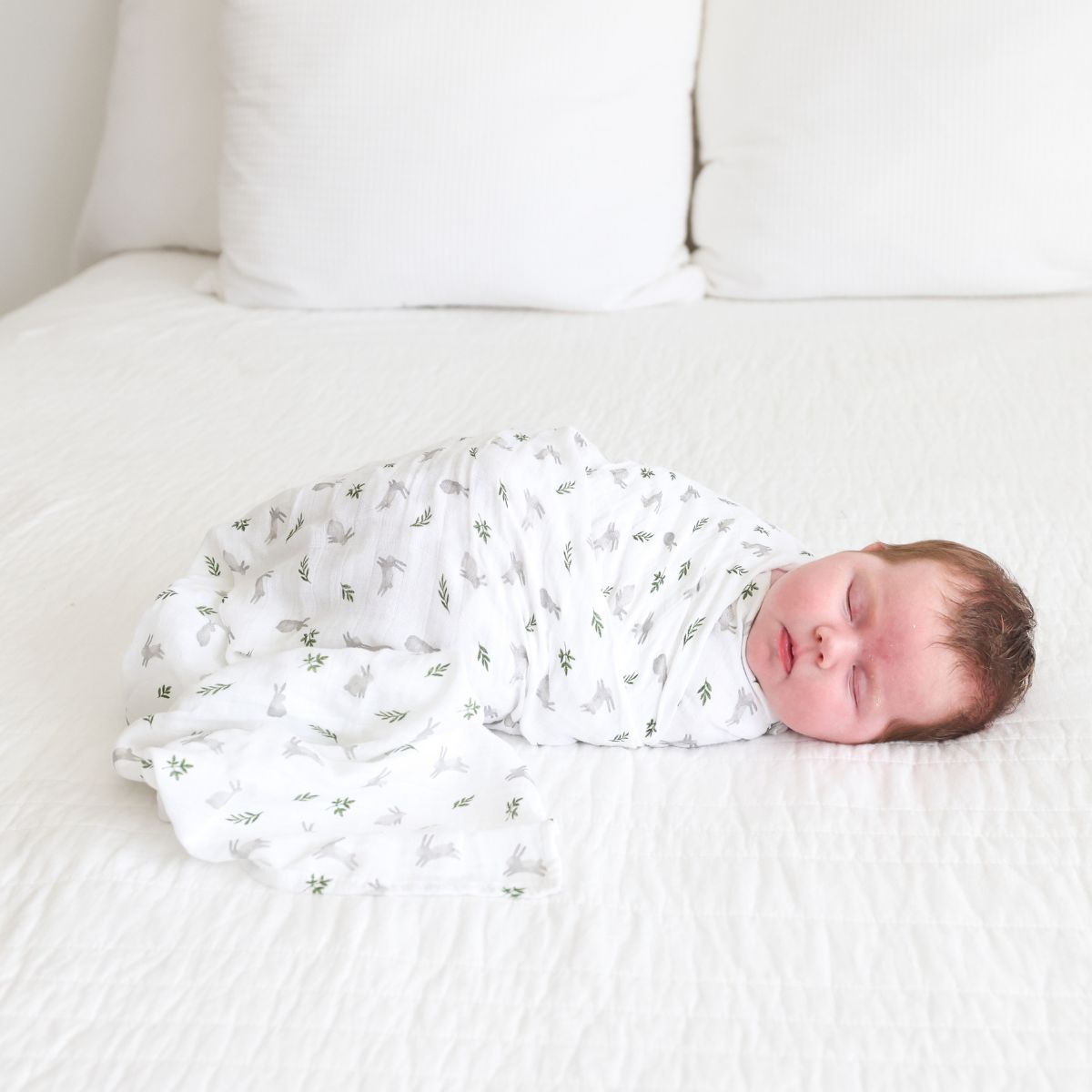 Newborn baby swaddled in a white blanket with green patterns, lying on a white bed.