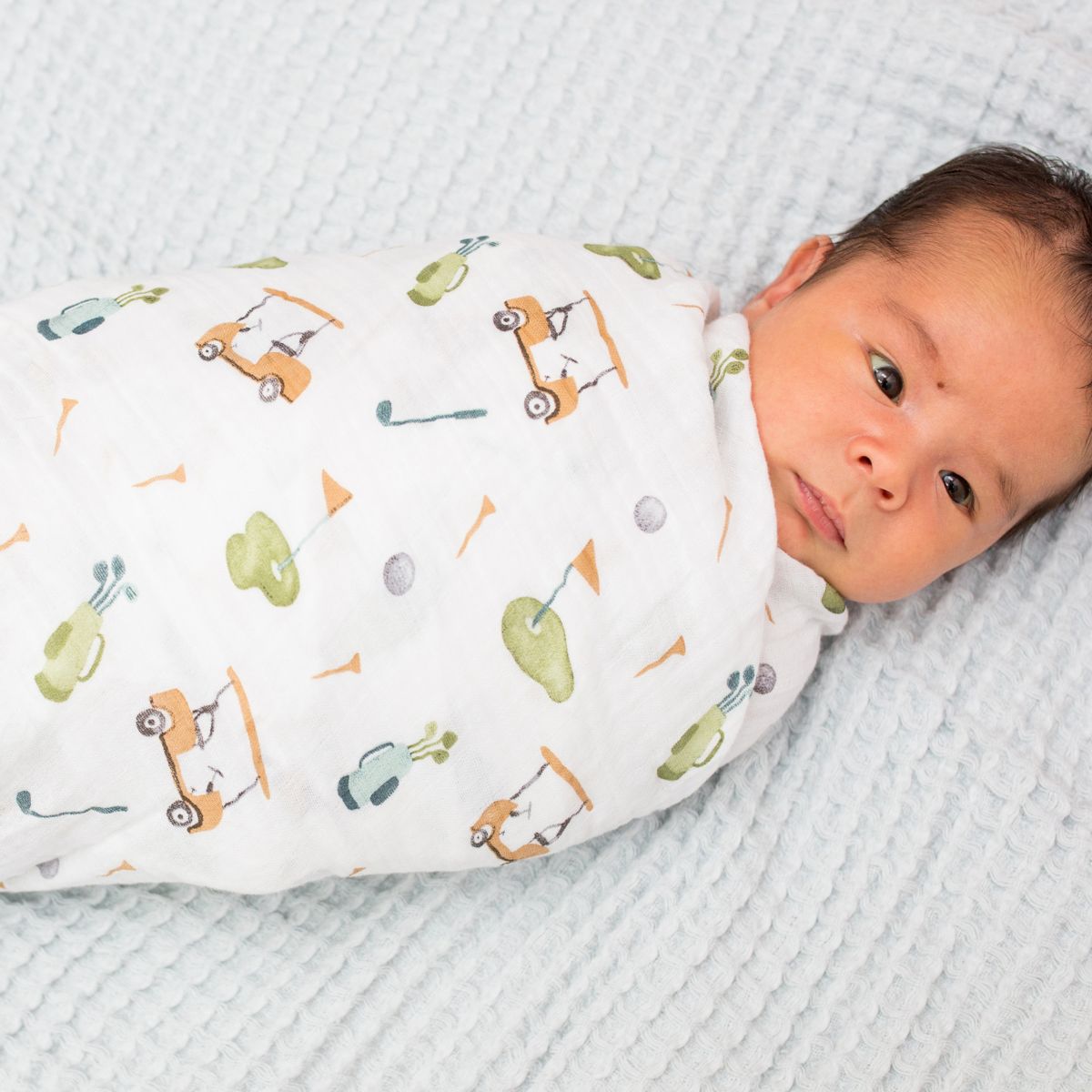 Newborn baby swaddled in a white blanket with golf-themed design on a textured surface.