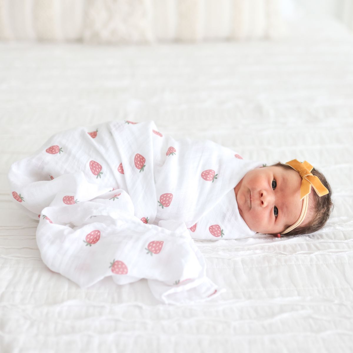 Newborn baby swaddled in a white blanket with pink patterns, wearing a yellow headband on a white background.