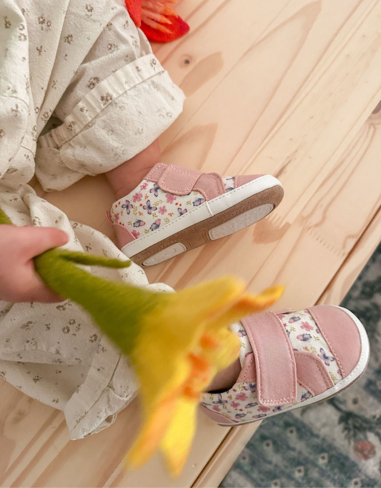 Child's feet wearing floral-patterned shoes on a wooden floor with a hand holding a yellow flower.