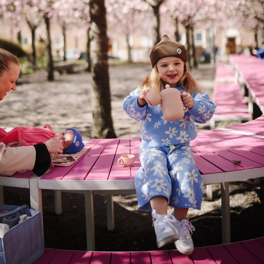 A young child wearing a blue floral print outfit and a brown beanie sits on a curved pink wooden bench outdoors, holding a small toy teapot, while another person tends to a baby nearby.