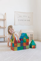 A young child playing on a rug, building castle structures with colorful translucent Magna-Tiles while a toy house sits in the background.
