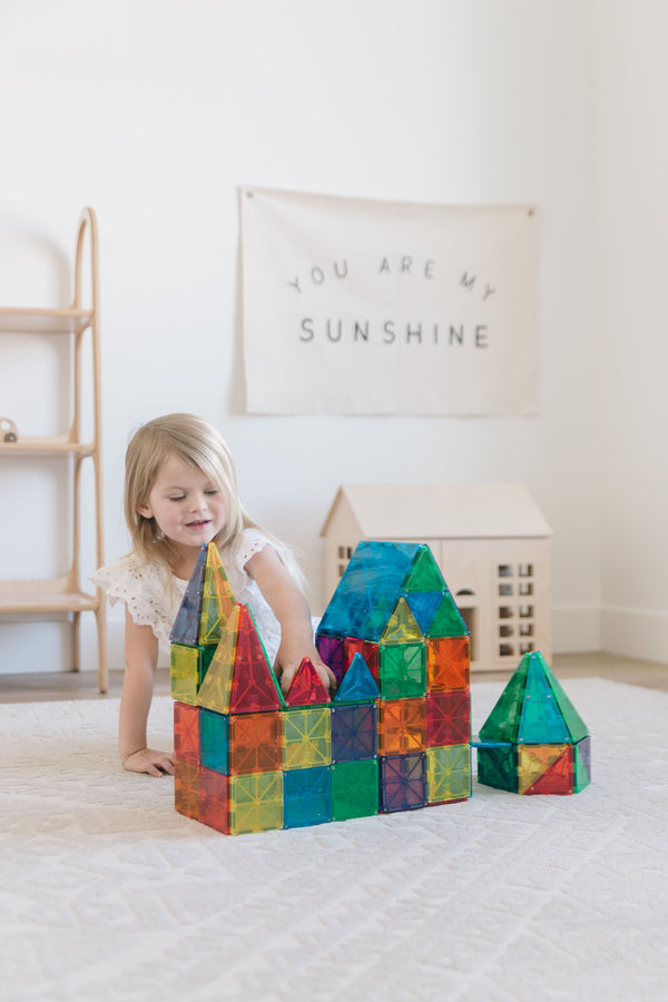 A young child playing on a rug, building castle structures with colorful translucent Magna-Tiles while a toy house sits in the background.