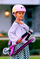 Child wearing a pink helmet and holding a purple scooter outdoors.
