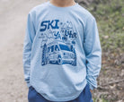 A child wearing a light blue long-sleeve t-shirt with a dark blue graphic print of a ski van, mountains, and the words 'SKI YA ISHER'.