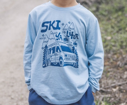 A child wearing a light blue long-sleeve t-shirt with a dark blue graphic print of a ski van, mountains, and the words 'SKI YA ISHER'.