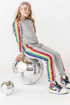 Person wearing a gray outfit with rainbow stripes sitting on a disco ball against a white background