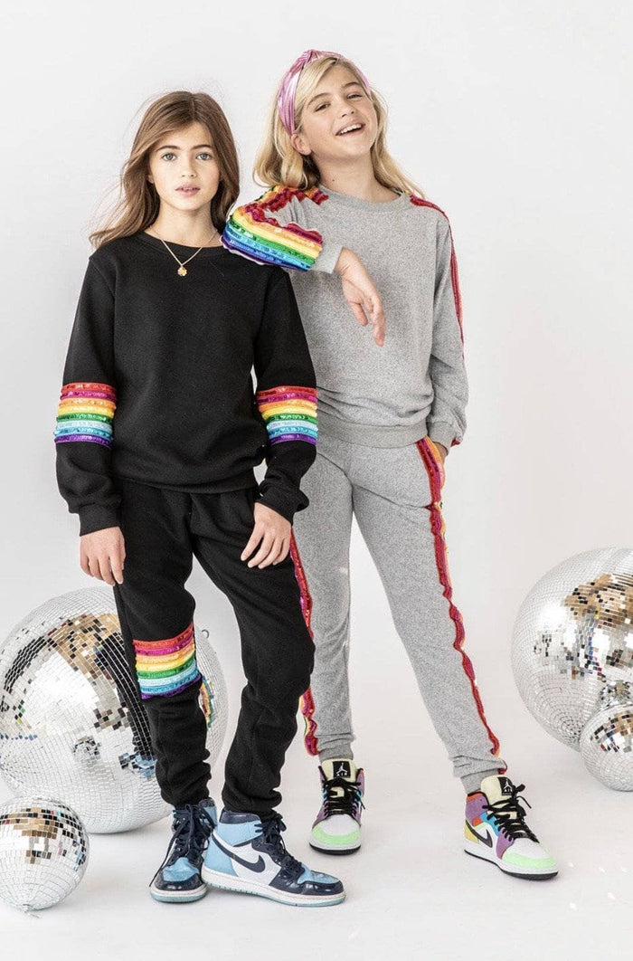 Two girls modeling matching rainbow-themed sweatshirt and jogger sets against a white studio background with disco ball props. One set is black with rainbow stripes on the sleeves and thighs, and the other is gray with red side stripes and rainbow shoulder detailing.
