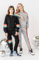 Two girls modeling matching rainbow-themed sweatshirt and jogger sets against a white studio background with disco ball props. One set is black with rainbow stripes on the sleeves and thighs, and the other is gray with red side stripes and rainbow shoulder detailing.