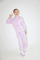 A young girl wearing a light purple velour tracksuit with colorful embroidered flowers, posing in a bright white room.