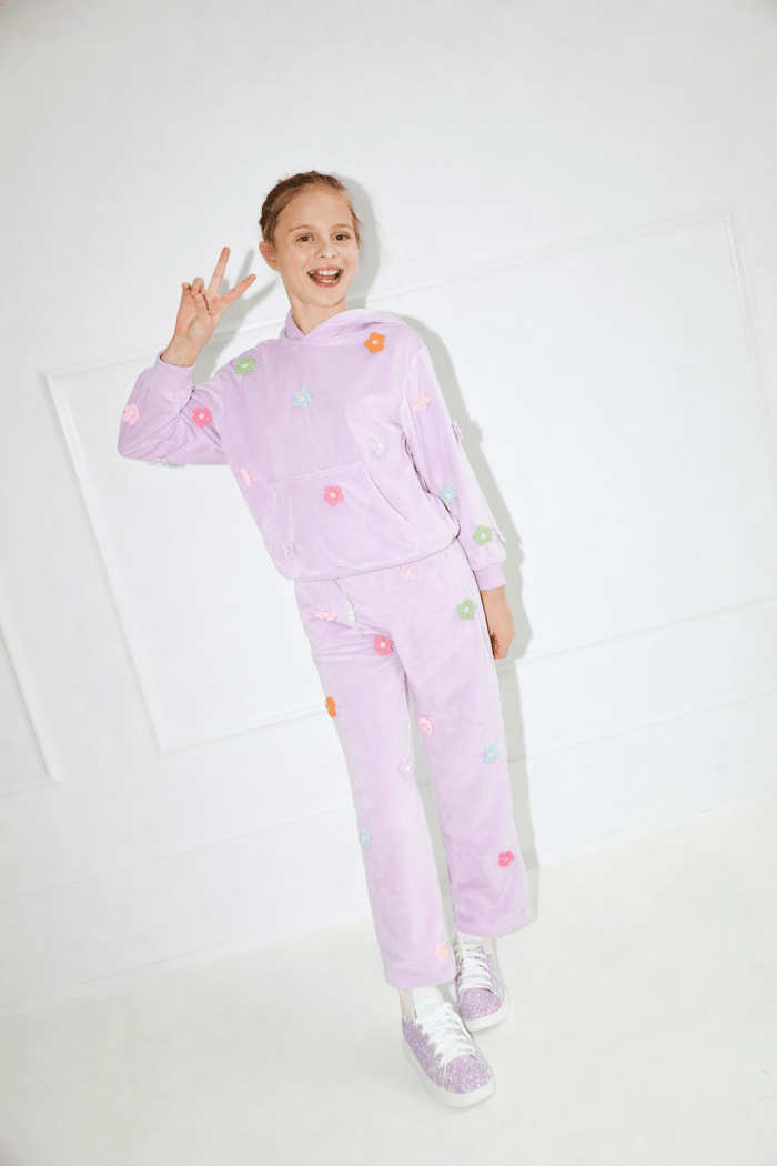A young girl wearing a light purple velour tracksuit with colorful embroidered flowers, posing in a bright white room.