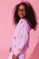 A young girl with curly dark hair, glasses, and a pink bow headband is wearing a light purple/lavender hooded sweatshirt and matching pants covered with colorful flower appliqués, posed against a solid pink studio background.