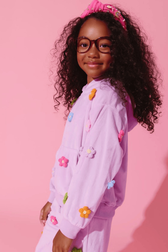 A young girl with curly dark hair, glasses, and a pink bow headband is wearing a light purple/lavender hooded sweatshirt and matching pants covered with colorful flower appliqués, posed against a solid pink studio background.