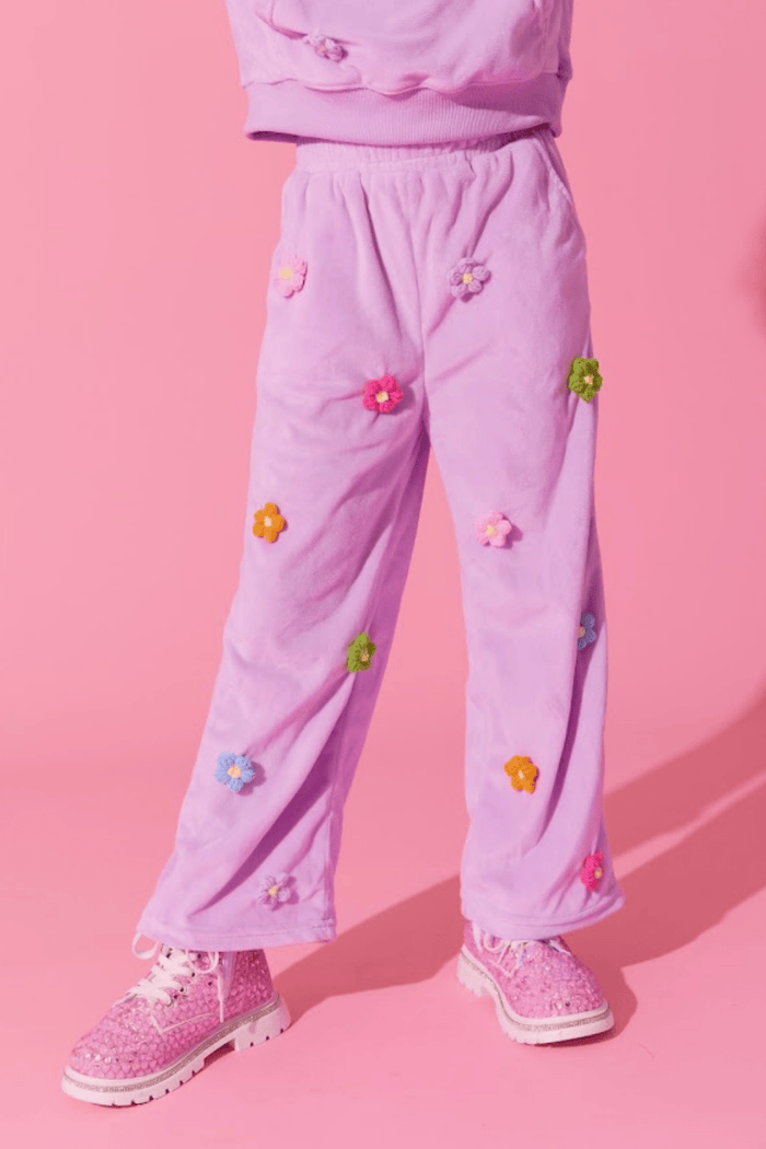 A child wearing lavender corduroy wide-leg pants with colorful embroidered flowers, paired with pink glitter sneakers, set against a solid pink background.