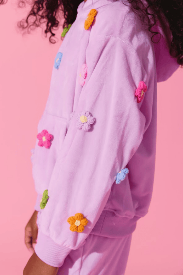 A close-up of a person wearing a lavender hooded sweatshirt with colorful crocheted flower appliqués in pink, orange, yellow, blue, and green, set against a solid pink background.