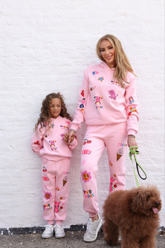 A woman and a young girl wearing matching pink graphic sweatpants and hoodies stand against a white wall, holding hands and accompanied by a brown dog on a leash. The matching outfits feature colorful cartoon ice cream cone and character prints.