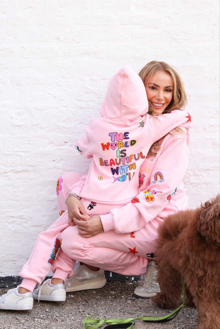 A woman and a child wearing matching pink embroidered hooded sweatshirt and jogger sets, hugging against a white brick wall, with a brown dog on a green leash in the foreground.