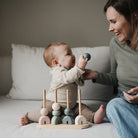 A toddler sitting on a white bed playing with a wooden multi-shape stacker toy while a woman sits beside them smiling.