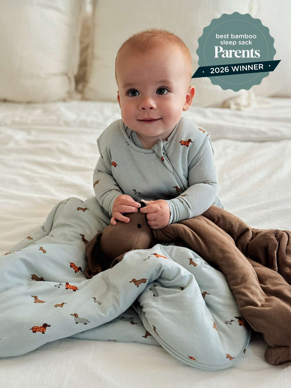 A baby sitting on a white bed wearing a light blue long-sleeve sleep sack with a brown dachshund print, wrapped in a brown plush blanket. A teal badge in the top right reads 'best bamboo sleep sack Parents 2026 WINNER'.