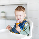 Child in a high chair wearing a blue bib, holding an ice cream cone.