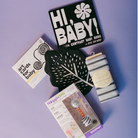 A baby gift set arranged on a light purple background, including a black 'HI, BABY!' book, 'art cards for baby' cards, a black leaf-shaped item, a striped 'Tinkle Blanket' in a box, and a NugginSilk sensory toy in its packaging.