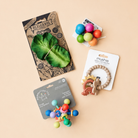Four baby accessories arranged on a warm beige background: a packaged green leaf teether from Olfic & Carol, a wooden bead teething ring with a rainbow and sun from mushie, a colorful sensory bead toy on a gray card, and a cluster of colorful round teething toys on a white card.