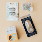 Four baby care products arranged on a warm beige background: a package of nanobebe pacifiers, a Natursüfteln box with a fox illustration, a packaged breast pacifier, and a small plastic bag of accessories.