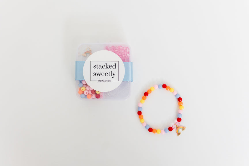 A clear plastic storage box labeled "stacked sweetly" containing pink and colorful beads, displayed alongside a finished stretchy bracelet featuring pastel and primary-colored beads with a gold rainbow charm against a white background.