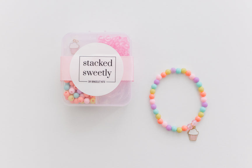A clear product photo of the Sweet Treat DIY Mini Bracelet Kit, featuring a sectioned plastic storage container filled with colorful pastel beads and a small cupcake charm, alongside a finished pastel-beaded stretch bracelet with a matching cupcake charm on a white background.