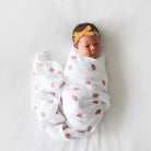 Newborn baby wrapped in a white blanket with red patterns, wearing a yellow headband on a light gray background.