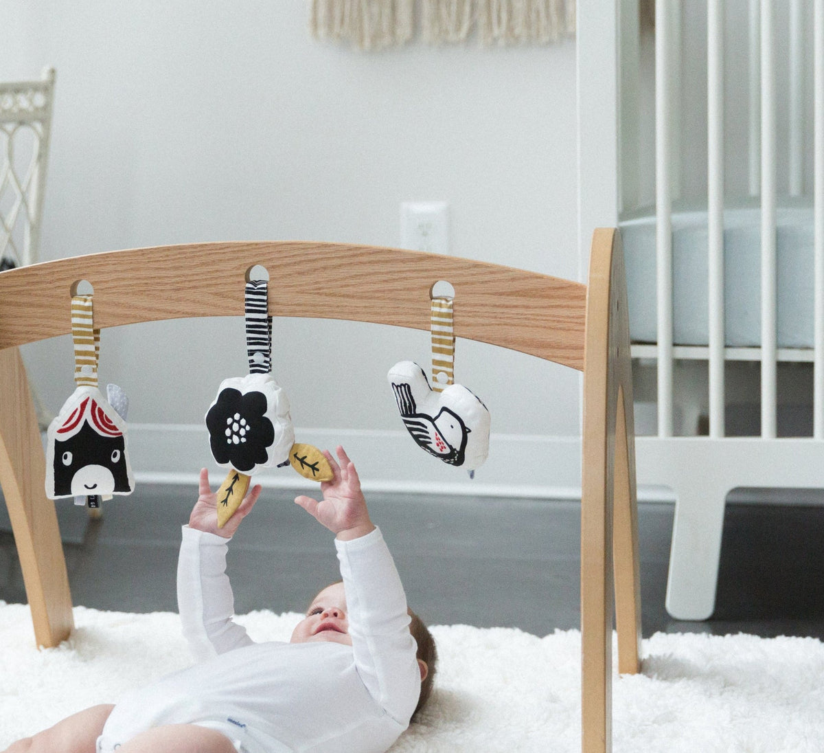 A plush Wee Gallery house stroller toy hanging from an activity gym next to other black-and-white toys, with a baby in the background.