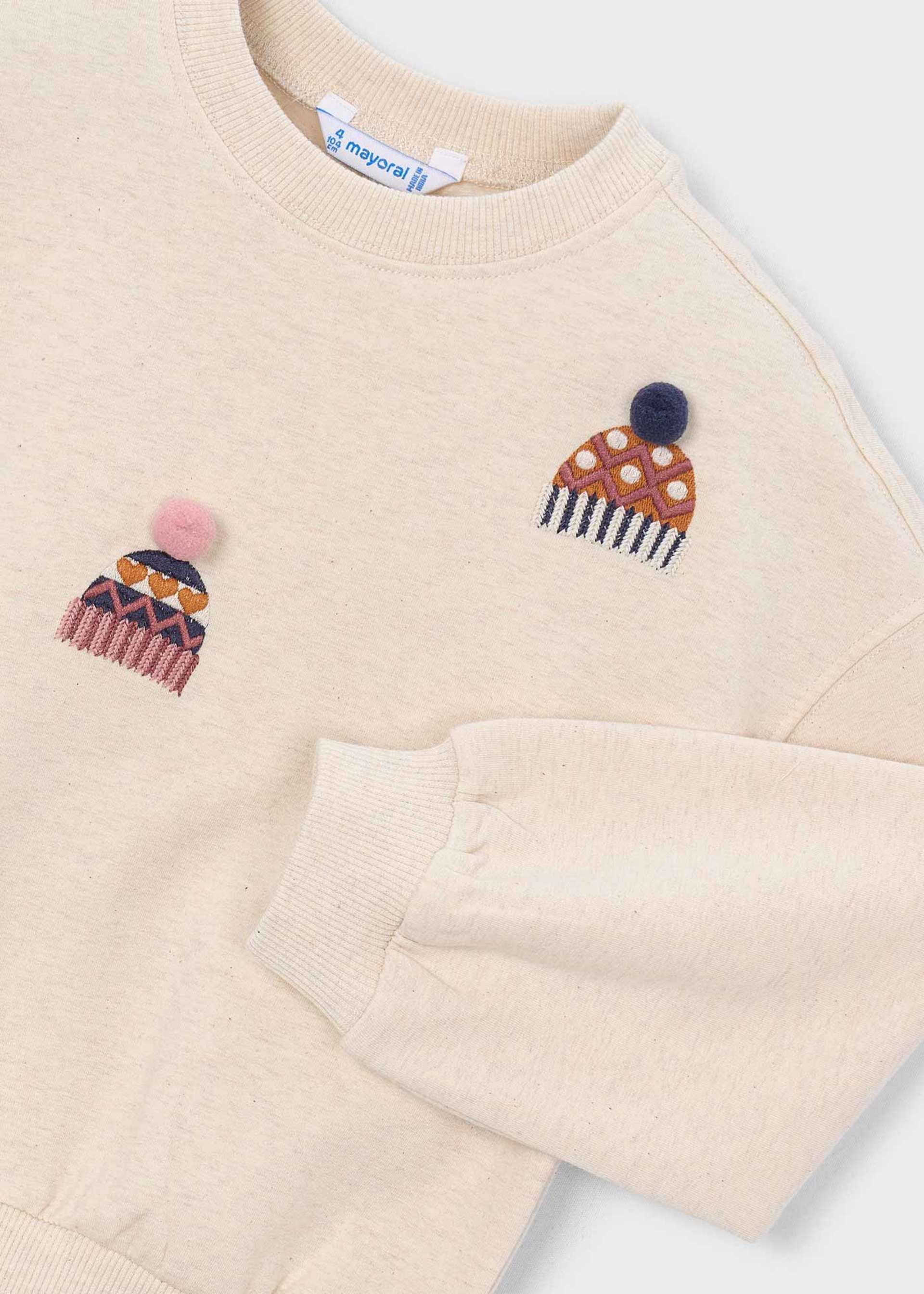 A close-up of a cream-colored crewneck sweatshirt featuring two embroidered knit hat motifs on the chest. The left chest has a pink and orange patterned hat with a pink pom-pom, and the right chest has a brown and orange polka-dot hat with a dark blue pom-pom.