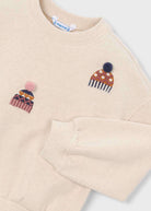 A close-up of a cream-colored crewneck sweatshirt featuring two embroidered knit hat motifs on the chest. The left chest has a pink and orange patterned hat with a pink pom-pom, and the right chest has a brown and orange polka-dot hat with a dark blue pom-pom.