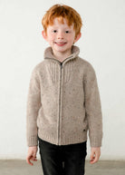 A young boy with red hair wearing a beige speckled knit zip-up sweater with a mock neck collar and black pants, standing against a plain off-white wall.