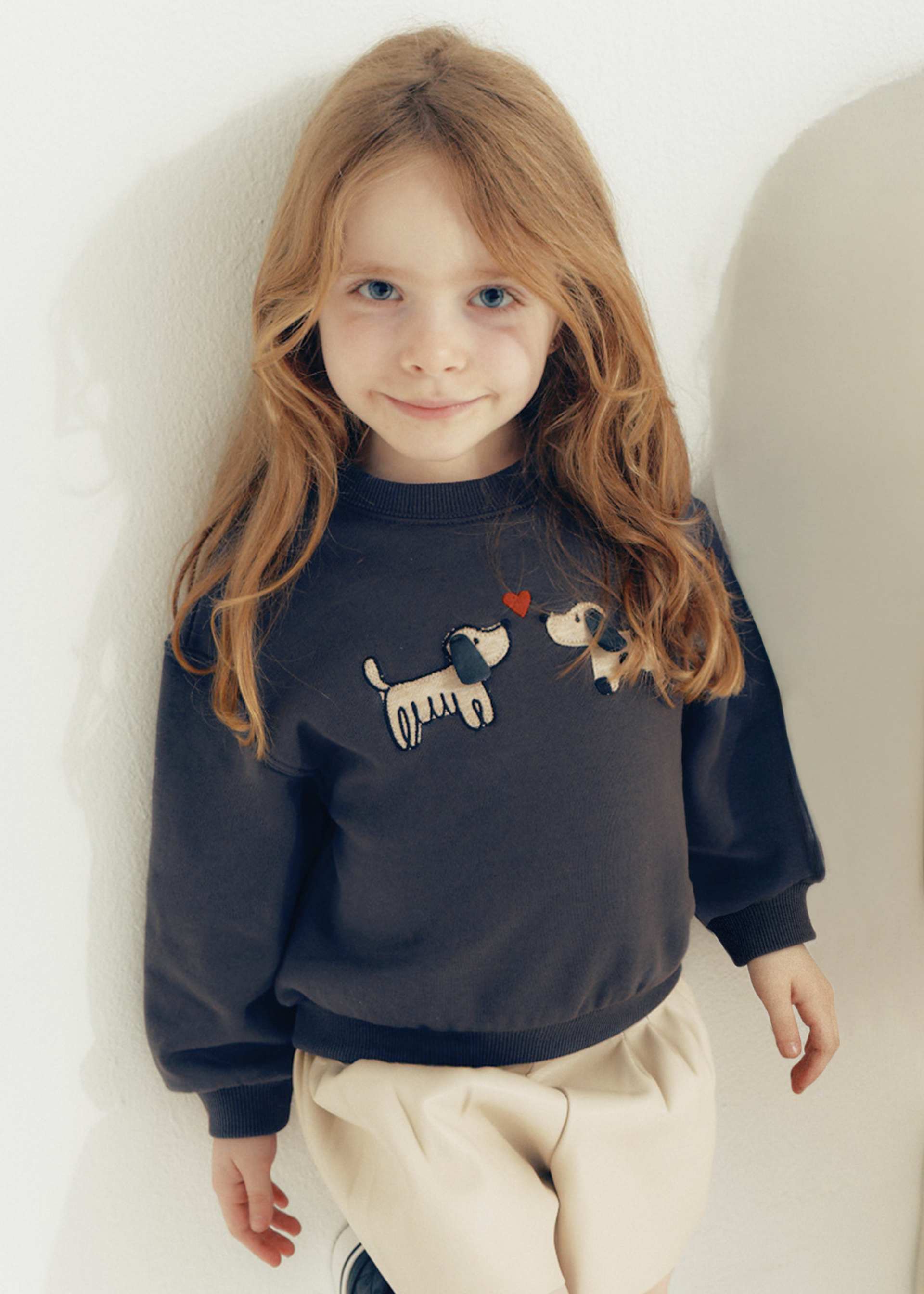 A young girl with long light brown hair wearing a dark charcoal gray sweatshirt with a dog graphic and cream-colored pants, posing against a plain off-white wall.
