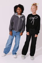 Two children modeling casual outfits against a plain light background. The child on the left wears a gray zip-up hoodie with blue jeans, and the child on the right wears a black long-sleeve top with a silver bow graphic and black patterned pants.