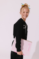 A young girl with blonde hair in a messy bun, wearing a black long-sleeve crop sweatshirt with a silver bow graphic and black leggings, holding a pink spiral notebook against a plain light background.