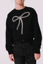 A person wearing a black cropped sweatshirt with a large sparkling silver bow graphic on the front, paired with black pants covered in small silver rhinestones, against a pale pink background.