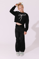 A young girl stands against a light pink studio background wearing a black cropped sweatshirt with a silver bow graphic, black pants covered in small silver rhinestones, and light green sneakers.