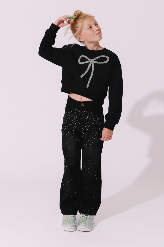 A young girl modeling a black long-sleeve crop top sweatshirt with a silver bow graphic, paired with black glittery pants and mint green sneakers, against a light pink studio background.