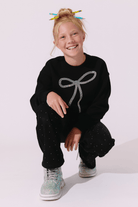 A young girl with blonde hair in a messy bun, decorated with colorful pencils, is crouching in a studio setting. She is wearing a black crewneck sweatshirt with a glittery silver bow graphic on the front, matching black glittery pants, and glittery sneakers.