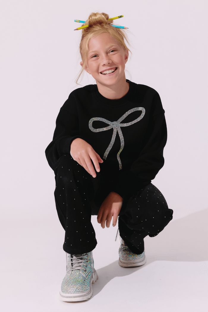 A young girl with blonde hair in a messy bun, decorated with colorful pencils, is crouching in a studio setting. She is wearing a black crewneck sweatshirt with a glittery silver bow graphic on the front, matching black glittery pants, and glittery sneakers.