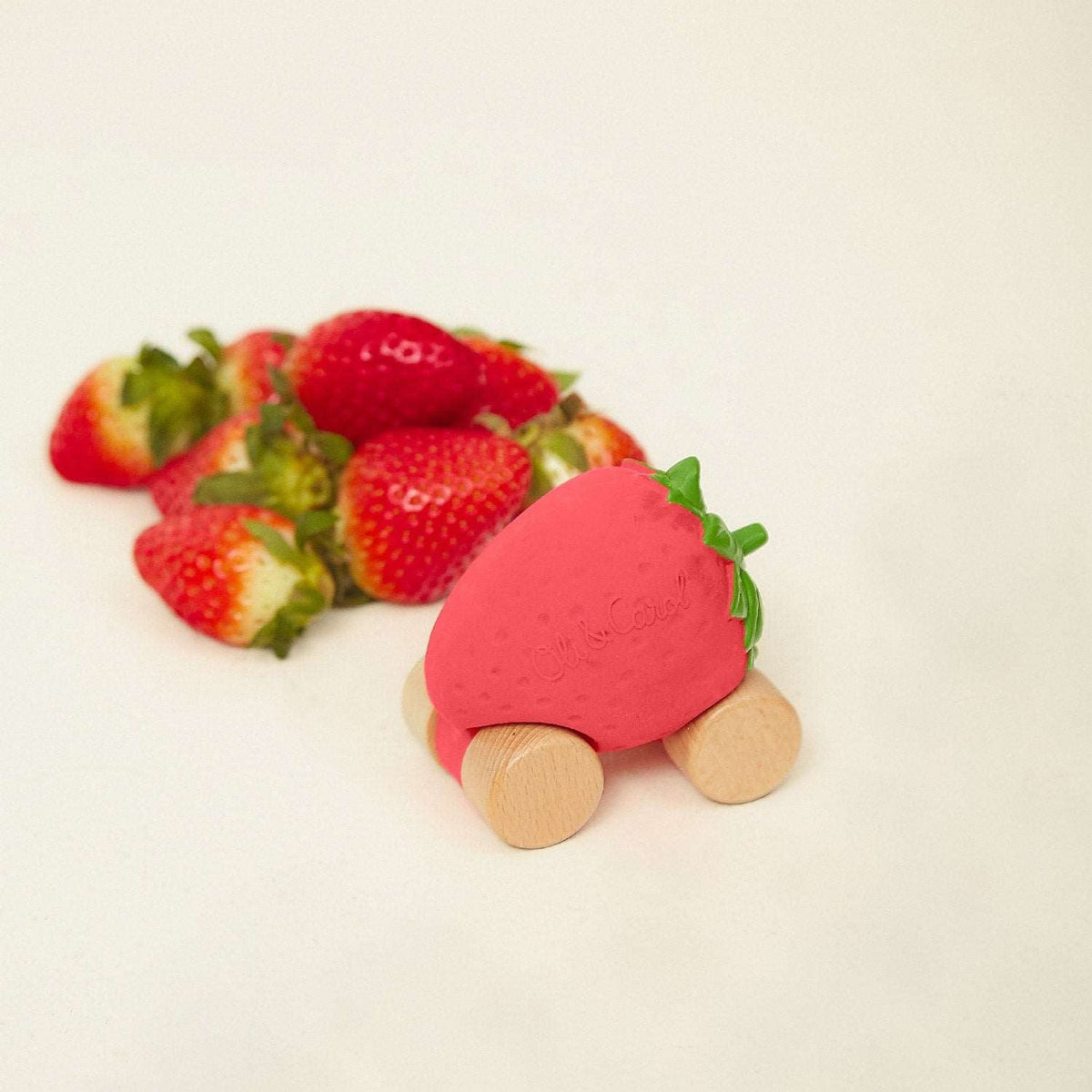 A strawberry-shaped wooden toy car alongside a cluster of fresh, ripe strawberries with green leaves.