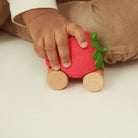 A toddler's hand holding a strawberry-shaped wooden toy car with a textured red body, green leafy top, and natural wood wheels.